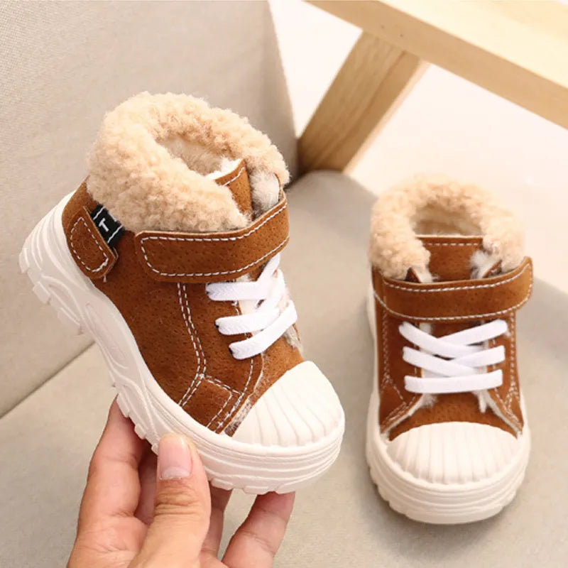 Main tenant la chaussure droite pour enfant de couleur marron.  La chaussure gauche est positionnée sur la chaise